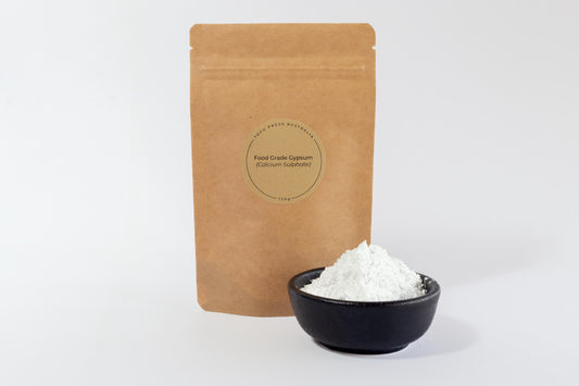 White food-grade gypsum powder (calcium sulphate) in a black bowl with a biodegradable, Kraft paper food pouch behind it on a white background.