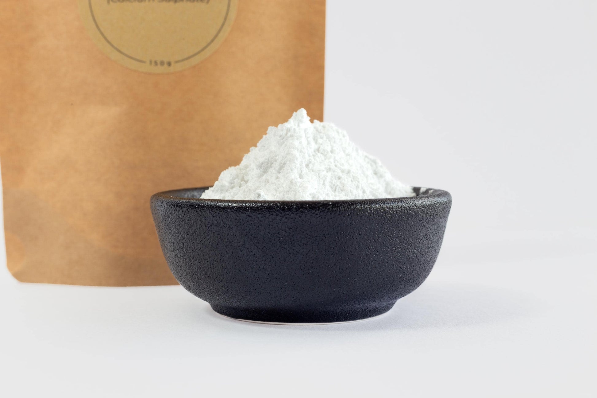Close up of white food-grade gypsum powder (calcium sulphate) in a black bowl with a biodegradable, kraft paper food pouch behind it.