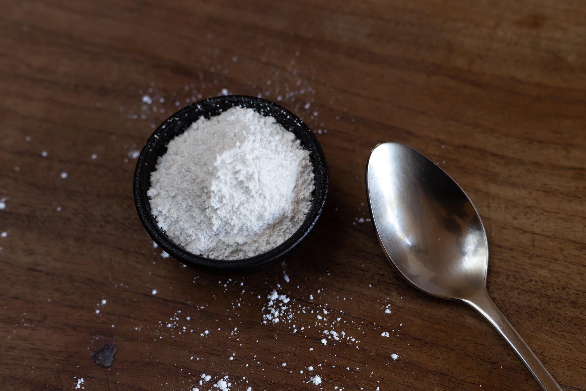 Close up of white food-grade gypsum powder in a small black bowl beside a metal spoon on a rustic wooden bench.